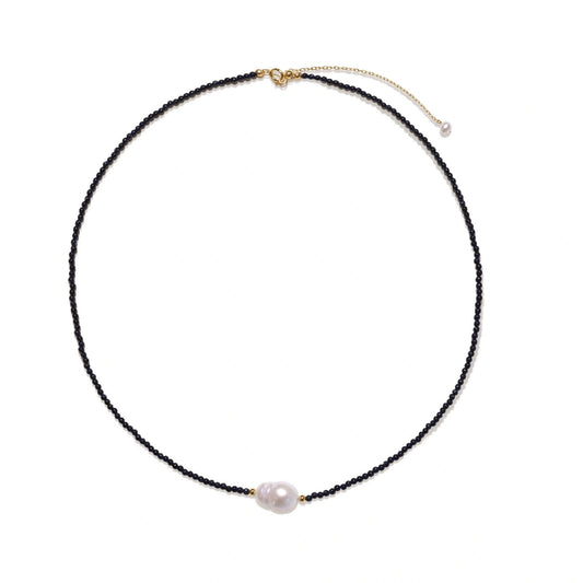 Delicate necklace with black blue sandstone beads and baroque pearl center on white background with gold accents.
